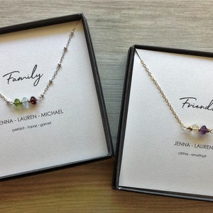 May include: Two gift boxes with necklaces inside. The box on the left has a silver chain with three gemstones and the text "Family" and "JENNA - LAUREN - MICHAEL peridot - topaz - garnet". The box on the right has a gold chain with two gemstones and the text "Friends" and "JENNA - LAUREN citrine - amethyst".
