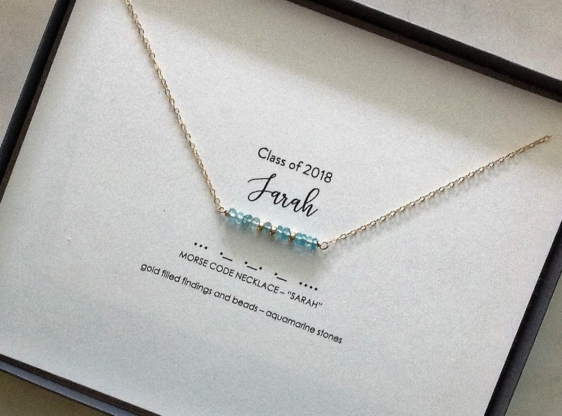 May include: A gold chain necklace with aquamarine stones spelling out "SARAH" in Morse code. The necklace is displayed in a white box with the text "Class of 2018" and "MORSE CODE NECKLACE - "SARAH"" printed on it.