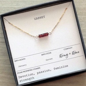 May include: A delicate gold chain necklace with a small red garnet gemstone bar pendant. The necklace comes in a box with the text "GARNET", "BIRTH MONTH January", "BIRTH STONE Garnet", "STONE PROPERTIES I POWERS Devotion, passion, feminine strength", "COMPANY String + stone".