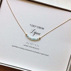 May include: A white card with the text "I DO" CREW Lynn, MORSE CODE NECKLACE, gold filled or sterling components and beads or glass beads". A gold chain necklace with blue and white beads is displayed on the card.