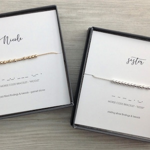 May include: Two gift boxes with delicate bracelets inside. The top box has a gold bracelet with the word "Nicole" spelled out in Morse code. The bottom box has a silver bracelet with the word "sister" spelled out in Morse code.