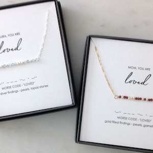 May include: Two necklaces in gift boxes. The top necklace is silver with blue and white beads and the text "LAURA, YOU ARE loved" and "MORSE CODE - "LOVED" sterling silver findings - pearls, topaz stones". The bottom necklace is gold with red and white beads and the text "MOM, YOU ARE loved" and "MORSE CODE - "LOVED" gold filled findings - pearls, garnet stones".