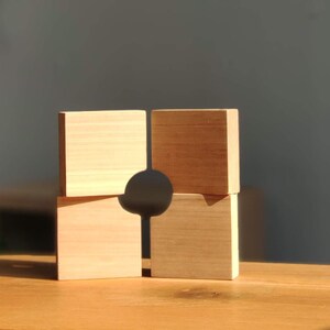 Puede incluir: Cuatro bloques de madera apilados en forma de cuadrado, con un agujero cortado en el centro. Los bloques son de color marrón claro y tienen un acabado liso y natural.