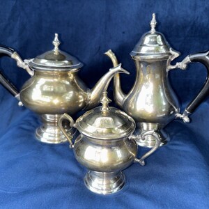 Puede incluir: Un juego de té de plata de tres piezas que incluye una tetera, una cafetera y un azucarero. Cada pieza presenta un acabado metálico pulido, una tapa con remate y una base de pedestal. La tetera y la cafetera tienen asas oscuras y curvas.