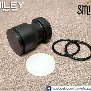 Puede incluir: Émbolo negro con un extremo circular plano, dos juntas tóricas negras y un disco circular blanco. La palabra "SMILEY" está en la parte superior de la imagen, junto con el texto "PLUNGER / CNC / POM". También hay un logotipo de carita sonriente.