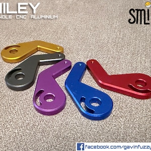 Puede incluir: Una variedad de manijas de pernos de aluminio CNC de colores dorado, gris, morado, azul y rojo. Cada manija tiene un diseño curvo único con una abertura circular. La palabra "SMILEY" se muestra en la imagen.