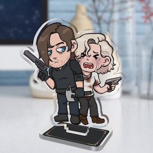 May include: A cartoon acrylic figure featuring two characters, one with a gun and the other crying. The figure is on a black base with gold accents. The characters are dressed in dark clothing and have detailed facial expressions.
