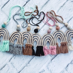 Może przedstawiać: Kolekcja odświeżaczy powietrza do samochodu w kształcie tęczy w różnych kolorach. Każda drewniana tęcza ma frędzel z makramy i sznur koralików. Tekst "SAWDUST JUNKEES" jest wydrukowany na każdej tęczy.