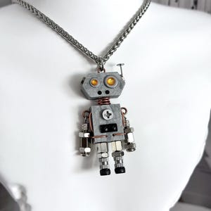 May include: A silver-toned robot pendant necklace featuring a hexagonal head with two amber-coloured circular eyes, a square torso with a central screw detail, and articulated limbs constructed from various metal nuts, bolts, and copper wire accents.