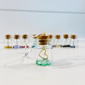 May include: A small glass bottle with a cork top and a gold heart charm hanging inside. The bottle is filled with green glitter and is attached to a white string.