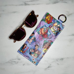 May include: A purple and blue patterned glasses case with a key ring. The case features a variety of Disney characters, including Ariel, Aladdin, Jasmine, and Simba.
