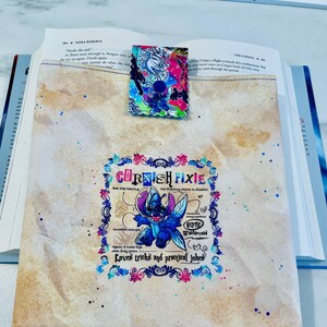 May include: A light brown book cover with a colorful illustration of a blue cartoon character with large ears and wings. The character is surrounded by flowers and vines. The text "CORNISH PIXIE" is written in a decorative font above the character. The text "Loves tricks and practical jokes" is written below the character.