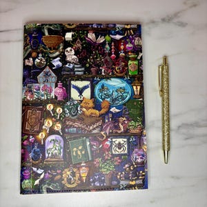 May include: A decorative notebook with a detailed, colorful illustration of a magical library scene, featuring books, potions, and a cat. A gold glitter pen rests beside the notebook, offering a writing tool. The notebook is a perfect gift for fantasy lovers.