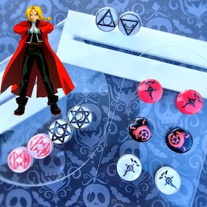 May include: A set of 10 small, round, clear glass earrings with various black and red symbols. The symbols include a star, a circle with a cross inside, and a circle with a triangle inside. The earrings are displayed on a black and white patterned surface.