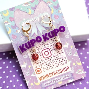 May include: Purple acrylic earrings with the text "KUPO KUPO" and red glitter ball charms. The earrings are displayed on a card with a whimsical design and the text "WHIMSYKEISHOP" and "whimsykei.com".