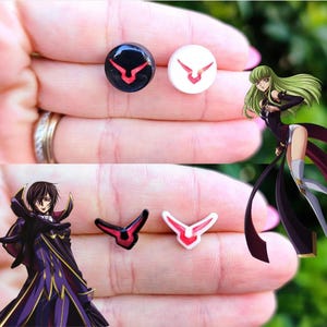 Puede incluir: Un par de pendientes negros y blancos con un símbolo rojo. El símbolo es una V estilizada con un punto en la parte superior y dos puntos en la parte inferior. Los pendientes se muestran en una mano.