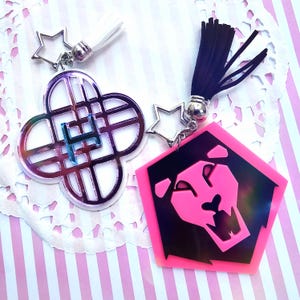 Puede incluir: Dos llaveros con cierres en forma de estrella. Uno tiene forma de trébol de cuatro hojas con un acabado iridiscente y una letra "H" azul. El otro es un pentágono rosa con un diseño de león negro y una borla negra.
