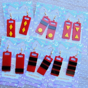 May include: Six pairs of rectangular red acrylic earrings displayed on individual light blue cards. The designs feature various yellow and black geometric accents, including diamonds, circles, triangles, and horizontal stripes, all attached to gold-tone hooks.