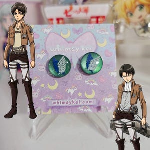 May include: A pair of round stud earrings with a green and blue design. Each earring features a white and blue wing emblem. The earrings are displayed on a purple card with the text "whimsy kei" and "whimsykei.com".