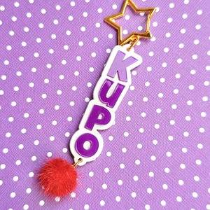 May include: Purple and white keychain with the name "KUPO" spelled vertically. The keychain has a gold star charm and a red fluffy pom-pom.