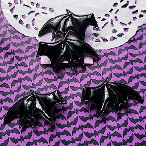 Puede incluir: Pinzas para el pelo negras con forma de murciélago y acabado brillante. Las pinzas están dispuestas sobre un fondo morado con un patrón de murciélagos repetido. Un tapete de encaje blanco está en el fondo.