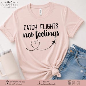 May include: A light pink short-sleeved t-shirt featuring the black text CATCH FLIGHTS not feelings, accompanied by a dashed line forming a heart shape and a small airplane icon. The shirt is displayed with blue denim jeans and a light-colored tumbler.