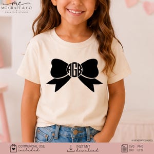 May include: A cream-colored short-sleeve t-shirt featuring a large black graphic of a bow. In the center of the bow is a circular monogram containing the letters AGB in a bold, black, sans-serif font. The shirt has a crew neckline.