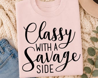 May include: A light pink crewneck sweatshirt featuring the black text Classy WITH A Savage SIDE printed on the front. The sweatshirt has long sleeves and a ribbed neckline, showcasing a bold script and sans-serif typography combination.