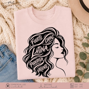 May include: A light pink crewneck sweatshirt featuring a black graphic of a profile silhouette with flowing hair. The hair contains the words Fuerte, Capaz, Latina, Poderosa, and Valiente in a stylized script font. The design is centered on the chest.