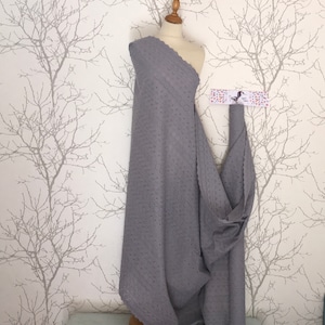 Puede incluir: Una tela gris calada drapeada sobre un maniquí. La tela tiene un borde festoneado y un patrón repetitivo de pequeños recortes circulares. La tela se exhibe contra una pared blanca con un diseño de ramas de árboles.