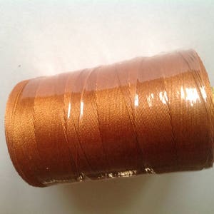 Puede incluir: Una bobina de hilo marrón envuelta en plástico transparente. El hilo está bien enrollado, mostrando una superficie texturizada. La bobina tiene un extremo amarillo. Adecuado para proyectos de costura y manualidades.
