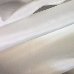 May include: Close-up of white fabric with subtle texture and folds. The fabric appears to be a light, possibly cotton or linen material, with a soft sheen. The folds create shadows and highlights, adding depth to the image.