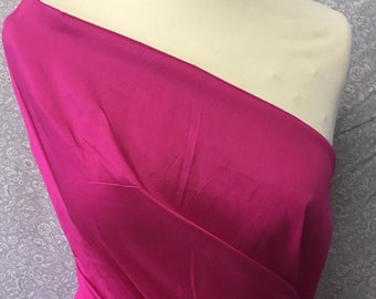 May include: A vibrant magenta fabric draped over a mannequin. The fabric has a smooth, lustrous appearance, with folds and shadows adding depth. The background is a light gray with a floral pattern.