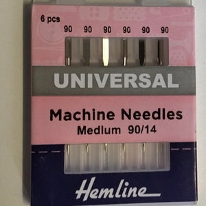 Könnte beinhalten: Eine Packung Hemline Universal Maschinennadeln, mittel 90/14. Die rosa-blaue Verpackung enthält sechs Nadeln, jede mit der Nummer 90 gekennzeichnet. Das Wort "UNIVERSAL" ist weiß auf grauem Hintergrund gedruckt. Die Packung hat auch den Text "6 pcs".