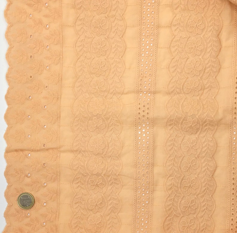 May include: Close-up of a peach-colored fabric with intricate eyelet and floral embroidery. The fabric features vertical stripes of detailed patterns, including scalloped edges and rows of small, evenly spaced holes. The design is symmetrical and detailed.