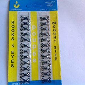 Peut inclure: Paquet de 24 crochets et oeillets noirs pour la couture. L'emballage jaune et bleu porte les inscriptions "HOOKS & EYES" et "poupée". Les crochets et oeillets sont en métal.