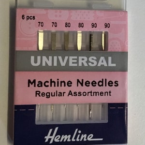 Könnte beinhalten: Eine Packung Hemline Universal-Maschinennadeln, reguläre Sortierung. Die rosa-marineblaue Verpackung enthält sechs Nadeln in den Größen 70, 70, 80, 80, 90 und 90. Die Nadeln sind in einem transparenten Kunststoffhalter sichtbar.