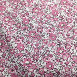 May include: A close-up of a fabric with a floral pattern. The fabric features a repeating design of pink flowers with white centres and green leaves on a white background. The flowers vary in size and shade of pink.
