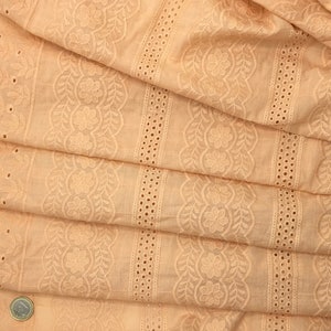 May include: Close-up of folded peach-colored fabric with intricate floral and leaf embroidery. The fabric features a scalloped edge and rows of eyelet detailing. The design includes a central floral pattern and vertical stripes with small circular cutouts.