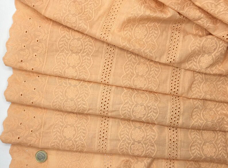 May include: Close-up of folded peach-colored fabric with intricate floral and leaf embroidery. The fabric features a scalloped edge and rows of eyelet detailing. The design includes a central floral pattern and vertical stripes with small circular cutouts.
