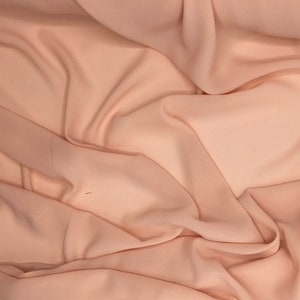 May include: A close-up shot of peach-colored fabric with soft folds and subtle shadows. The fabric appears to be lightweight and flowing, with a smooth texture. The overall effect is one of gentle movement and a delicate appearance.