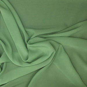 May include: A close-up shot of a flowing fabric in a soft, muted green hue. The material is draped and folded, creating gentle shadows and highlights. The fabric appears to be lightweight and smooth, suggesting a delicate texture.