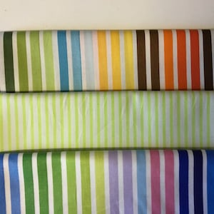 Puede incluir: Tres rollos de tela con rayas verticales en varios colores. El rollo superior presenta rayas marrones, verdes, azules, amarillas y naranjas. El rollo del medio tiene rayas verde claro. El rollo inferior tiene rayas azules, verdes, moradas y rosas.