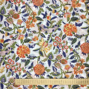 May include: White fabric with a repeating floral pattern. The design features orange, yellow, and red flowers, green leaves, and blue accents. A ruler is placed across the bottom of the fabric.