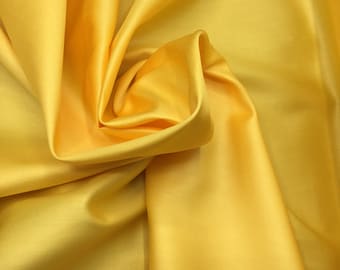 Pode incluir: Um tecido amarelo vibrante com uma textura de superfície lisa e lustrosa. O material está drapeado em dobras suaves e fluidas que criam sombras profundas e reflexos brilhantes, destacando a cor sólida e o brilho característico do tecido.