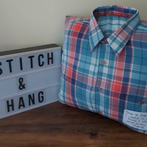 May include: A blue and red checked shirt pillow with the text "This is the shirt I used to wear hold it close and know I'm there" on the bottom right corner. The pillow is sitting on a wooden surface with a light blue sign that says "@STITCH & HANG" in black letters.