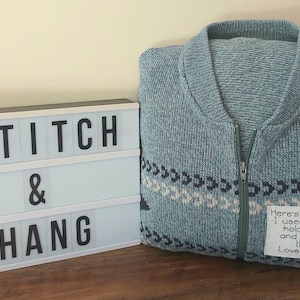 May include: A blue knitted cardigan with a white and black patterned design. The cardigan has a zipper and a handwritten note attached to it that reads "Here's the cardie I used to wear hold it close and know I'm there. Love Ray xXx".