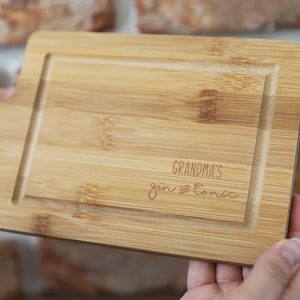 May include: A natural wood chopping board with a handle. The board has a groove around the edge and the text "Grandma's Gin & Tonic" is engraved on the surface.