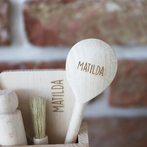 May include: A collection of wooden kitchen utensils, including a spoon, brush, and spatula, all engraved with the name "MATILDA." The items are displayed in a wooden container against a brick wall background. The utensils are made of light-colored wood.