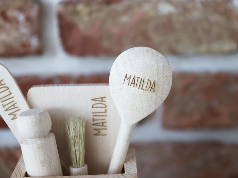 May include: A collection of wooden kitchen utensils, including a spoon, brush, and spatula, all engraved with the name "MATILDA." The items are displayed in a wooden container against a brick wall background. The utensils are made of light-colored wood.